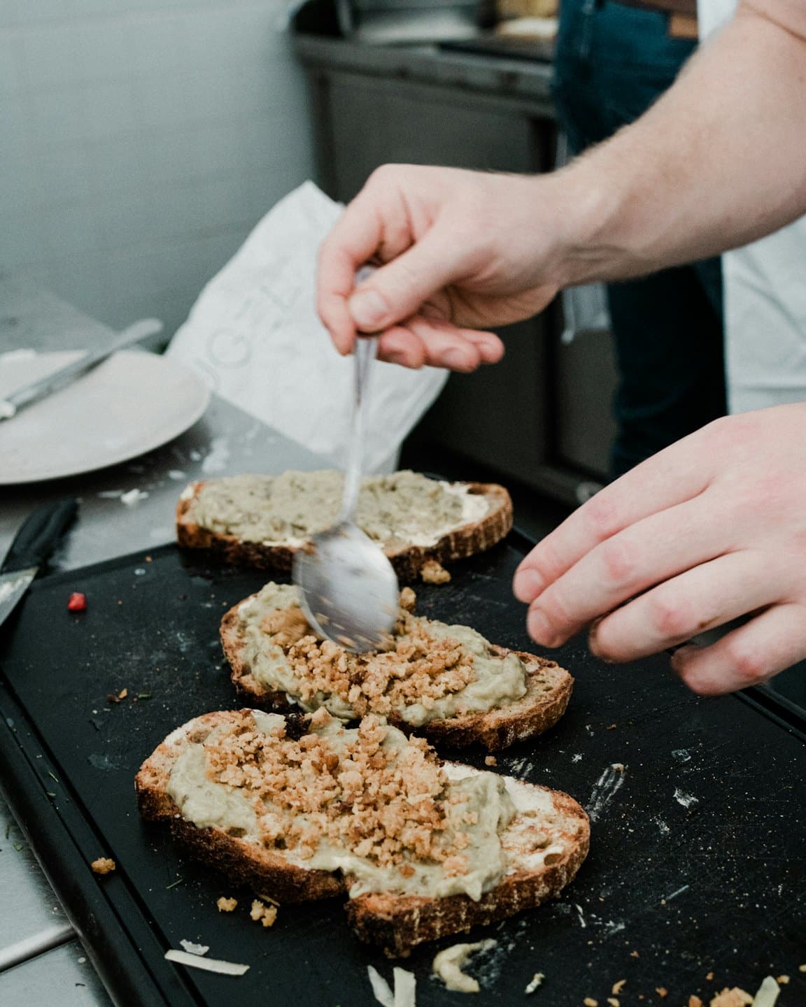 Hands spread green paste and topping onto slices of toasted bread.