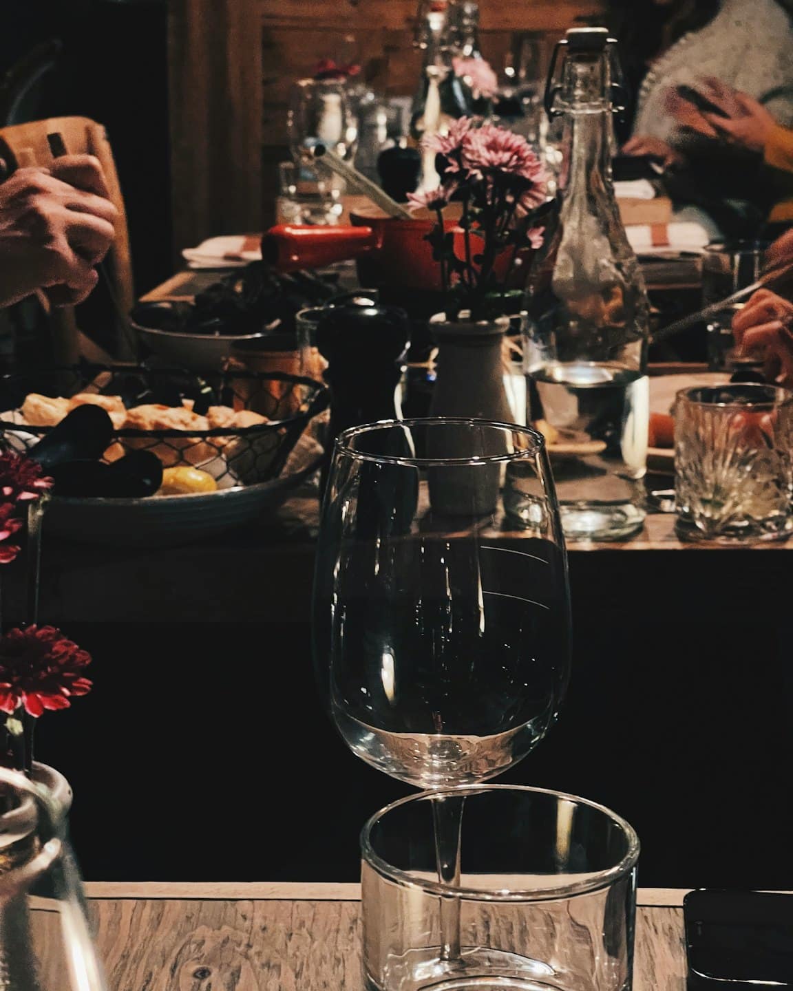 A wine glass sits on a wooden table with blurred hands reaching for food.
