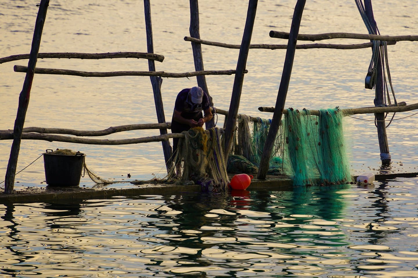 Man mends fishing nets on a wooden platform over water.