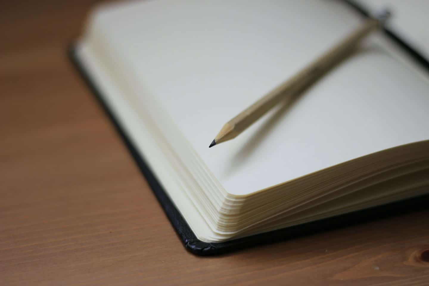 A pencil rests on an open, blank notebook on a wooden desk.