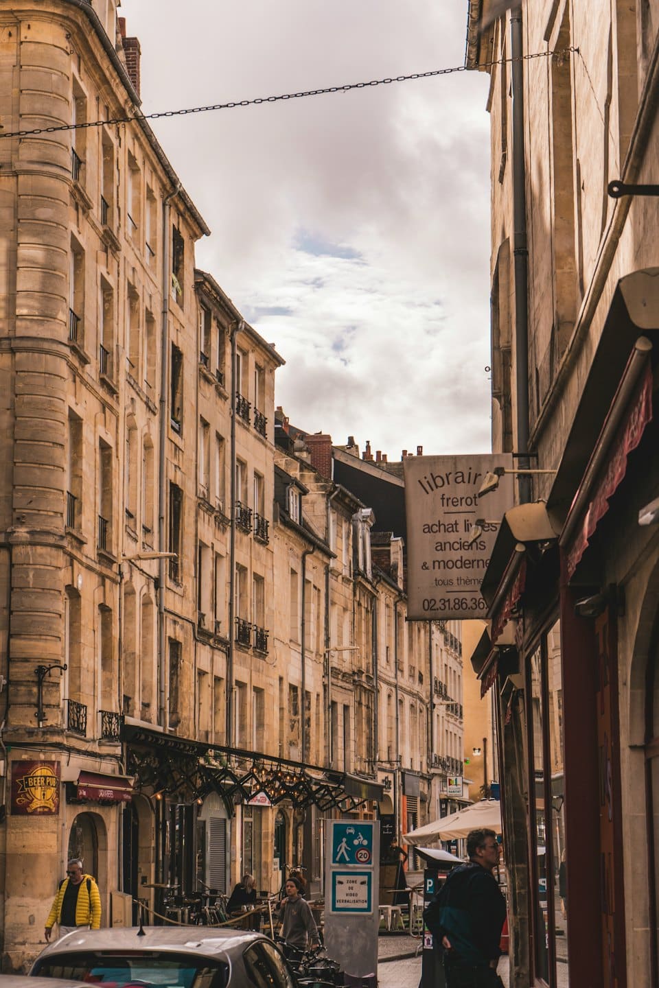 Pedestrians walk past shops along a narrow European stone street.