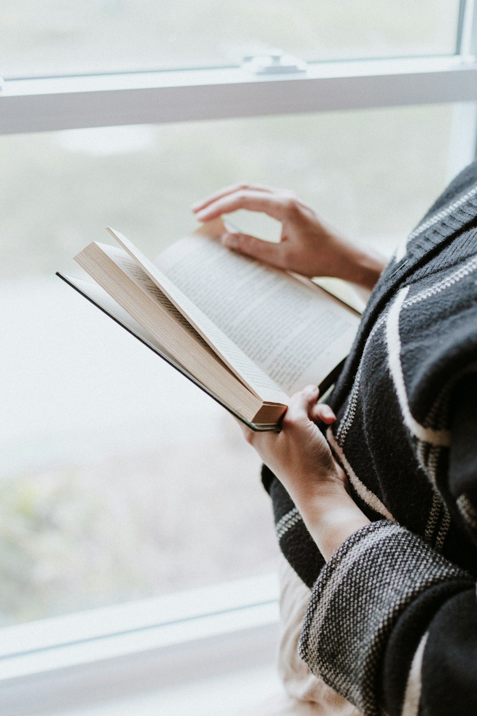 A person holds an open book while reading near a window.