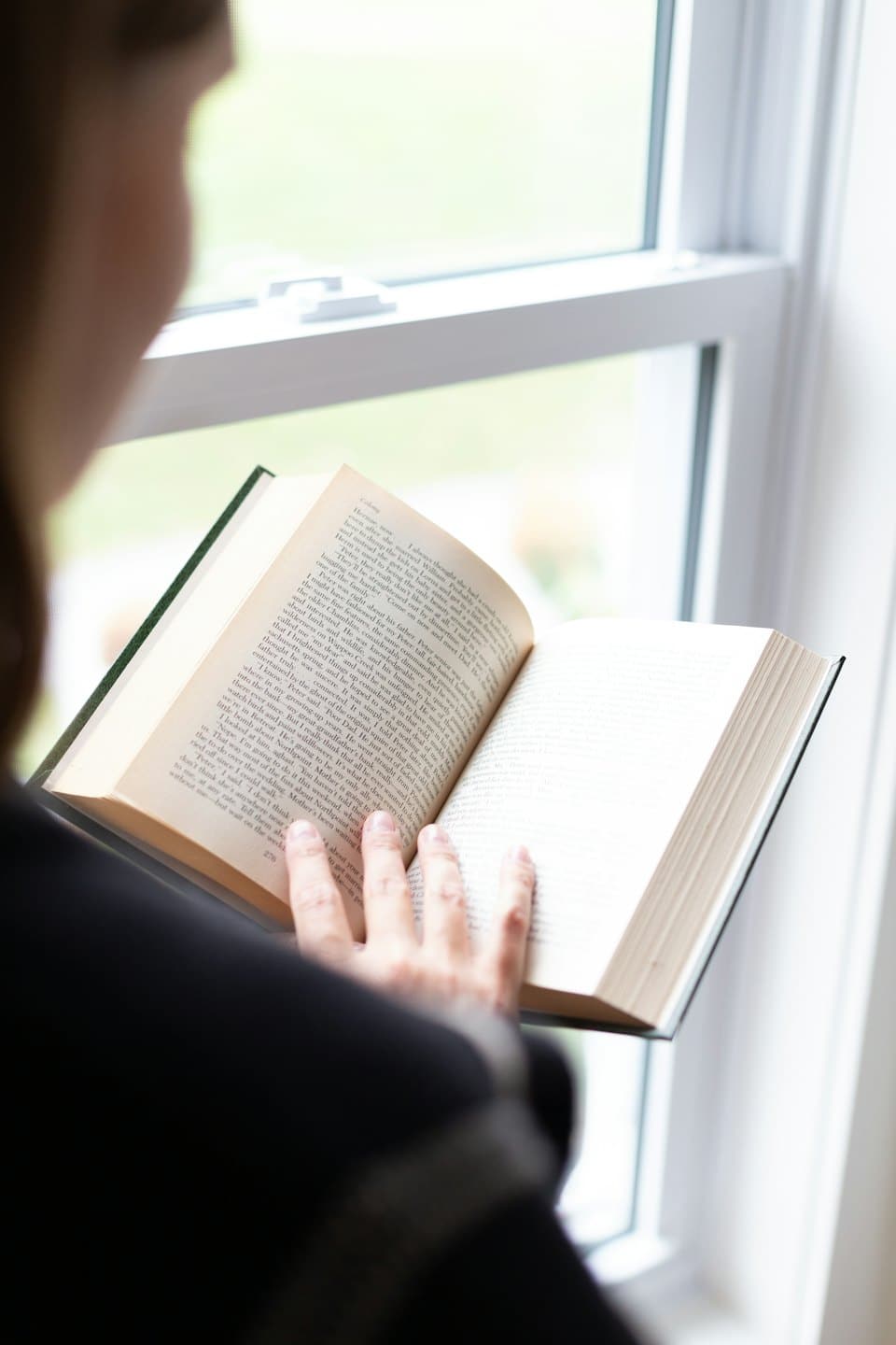 Person reading an open book near a bright window.